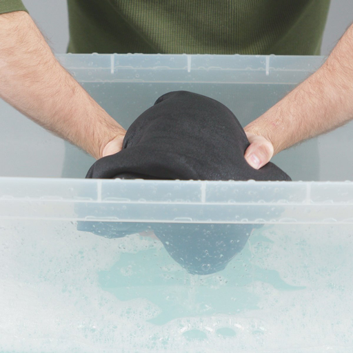 Hand model about to hand wash neoprene suit in a transparent basin with Gear Aid Revivex Wetsiut Shampoo diluted in water